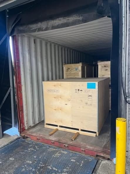 A large plywood crate stencilled NOVAIR standing on a pallet at the mouth of a shipping container on a loading dock.