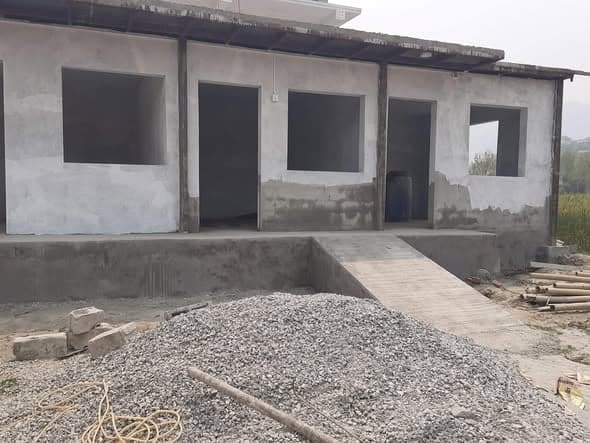 A newly built single-storey block with rendered walls, open window and door frames, a concrete ramp at the entrance and gravel piled in front.