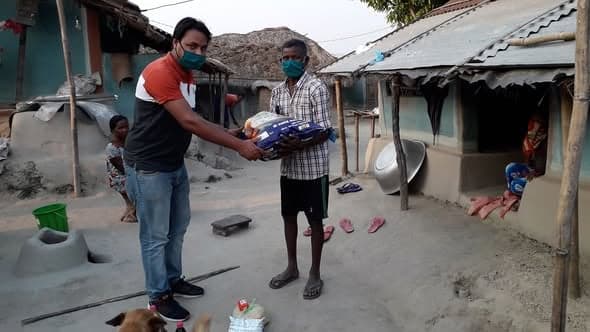 A man in a face mask hands a large sack of rice to a barefoot man outside mud-walled houses while a woman watches from a doorway.