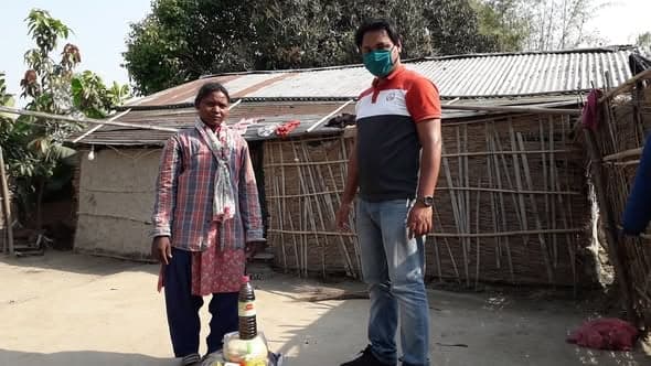 A woman in a checked shawl and a masked volunteer stand beside a bundle of supplies in front of a house walled with woven bamboo.