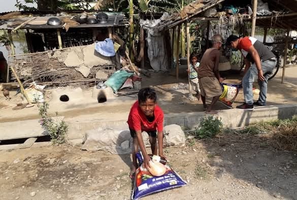 A boy in a red shirt crouches to lift a sack of rice and a bottle of oil outside a bamboo and thatch home while people gather behind him.