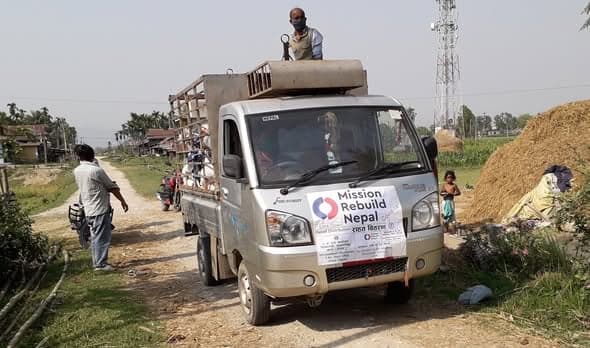 A small pickup truck carrying supplies, a Mission Rebuild Nepal relief notice fixed to its front, driving along a dirt village road.
