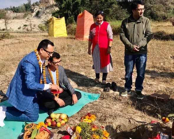 Two men in marigold garlands sit cross-legged on a mat on cleared ground, making offerings over brass plates of fruit and flowers while others stand watching.
