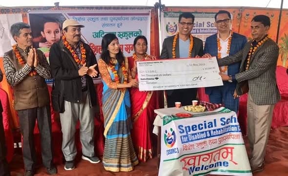 A line of people in marigold garlands hold up an oversized cheque made out to SSDRC for ten thousand US dollars, in front of a Special School banner.