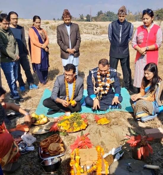 A crowd stands around two men seated on a mat for a foundation ceremony, with plates of offerings, flowers and a small shrine set on freshly dug earth.
