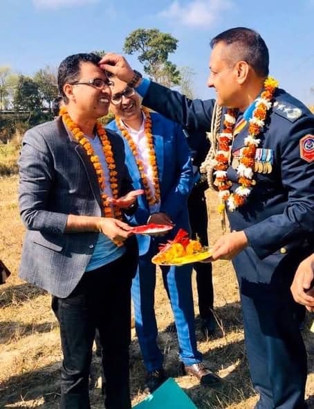 A man in a garland applies tika to the forehead of a uniformed officer while another guest looks on, all wearing marigold garlands.