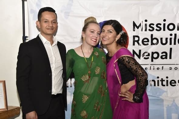 Three people in formal and traditional dress stand together in front of a Mission Rebuild Nepal banner.