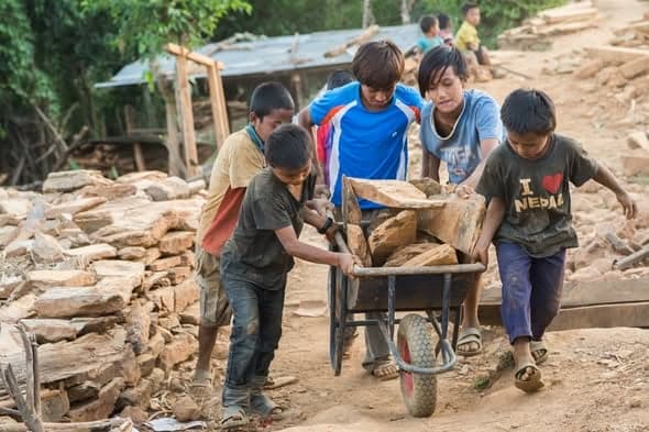 Five boys push a wheelbarrow loaded with cut stone along a dirt track past stacks of building stone.