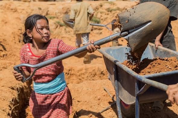 A girl in a red checked dress swings a long-handled shovel to load earth into a wheelbarrow on a bare construction site.