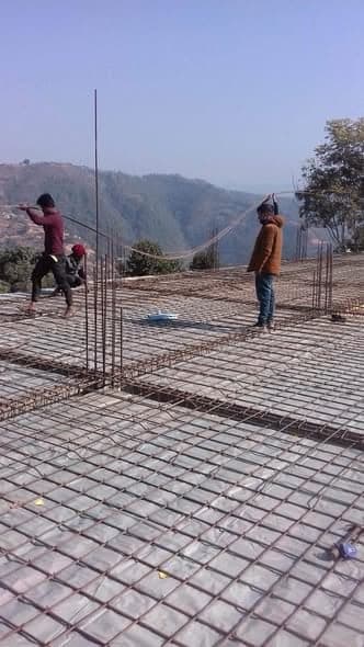 Two men tie steel reinforcing bars on a slab under a clear sky, the hills of Sindhupalchowk behind them.