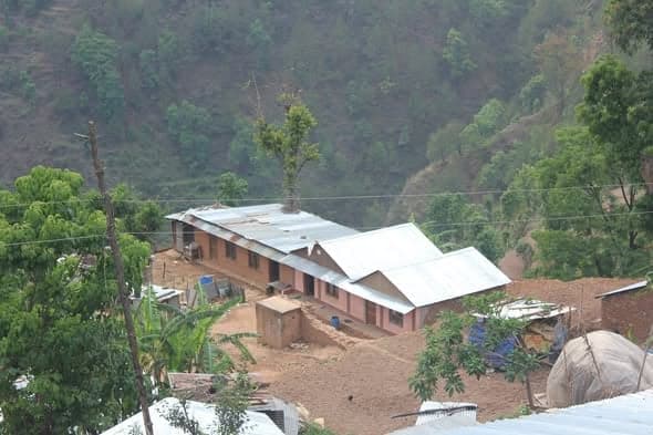 Two long buildings with new corrugated roofs stand together on a cleared terrace surrounded by forest.