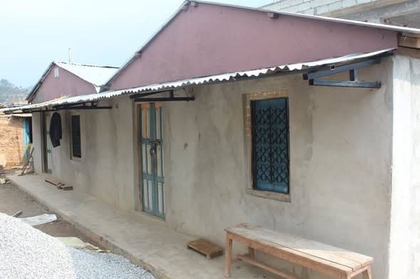 A plastered single-storey house with a deep porch roof, a green door and a wooden bench outside.