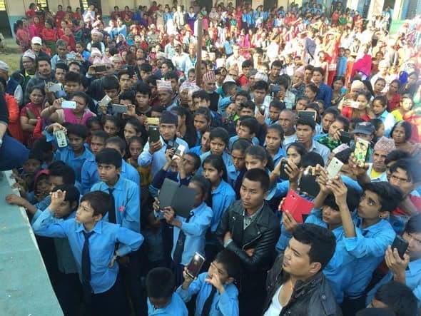 A dense crowd of schoolchildren in blue uniforms and villagers hold up mobile phones to photograph something out of frame.