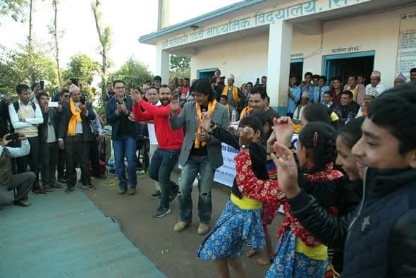 Girls in traditional dress dance on a concrete forecourt while men clap and film them outside a school building.