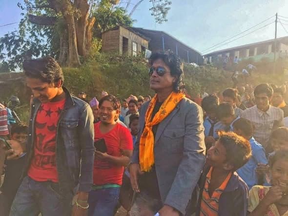 A man in a grey jacket and orange scarf walks through a press of villagers and children on a slope.