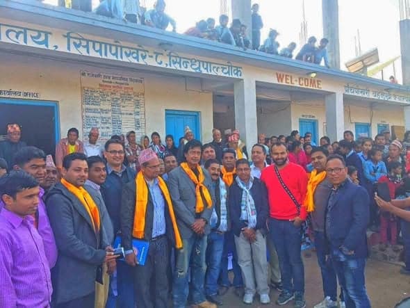 A line of guests in yellow scarves stand for a photograph outside a school office, with people crowded on the roof behind them.