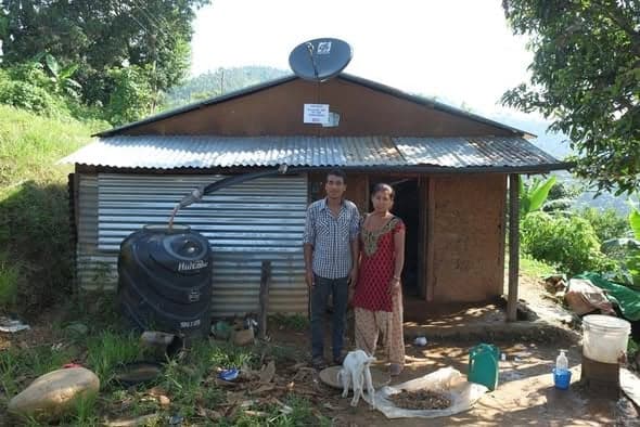 A man and a woman stand outside a corrugated-iron house with a satellite dish on the roof and a black water tank at the side; a goat stands in the yard.