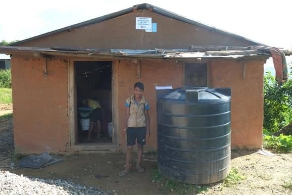 A boy stands in the doorway of a mud-walled house with a metal roof and a large black water tank beside it.