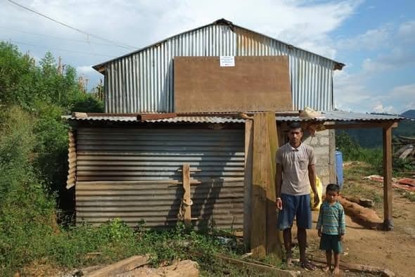 A man and a small boy stand at the corner of a corrugated-iron house raised on timber posts on a slope.