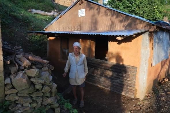An elderly man with a walking stick stands outside a mud-plastered house beside a stack of firewood and cut stone.