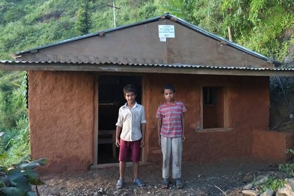 Two boys stand side by side in front of a mud-walled house with a corrugated-iron roof.