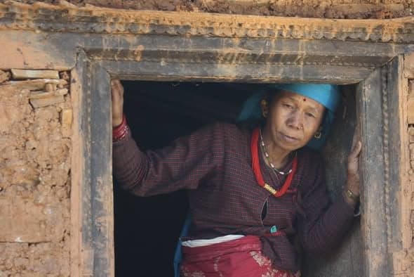 A woman in a red patterned blouse and blue headscarf leans out of the timber-framed doorway of an old mud house.