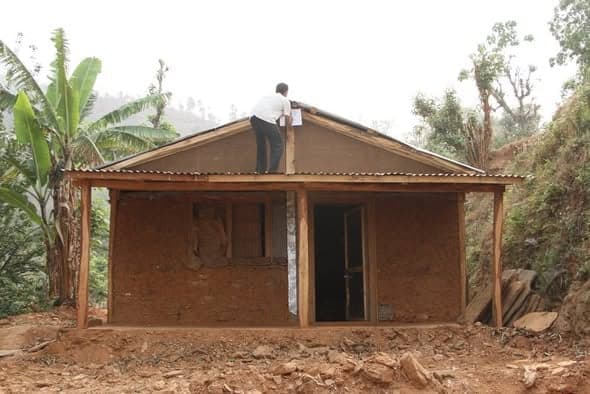 A man kneels on the ridge of a mud-walled house fixing corrugated sheets, banana trees rising behind him.