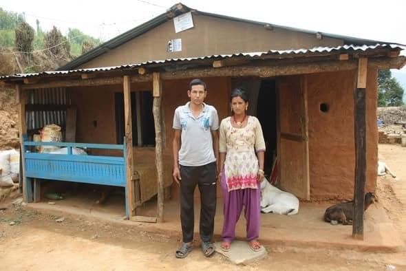 A young couple stand in front of a mud house with an open veranda, a blue wooden bench and a calf lying in the shade.