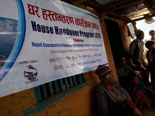A House Handover Program 2016 banner hangs on a wall while villagers sit beneath it in the shade.
