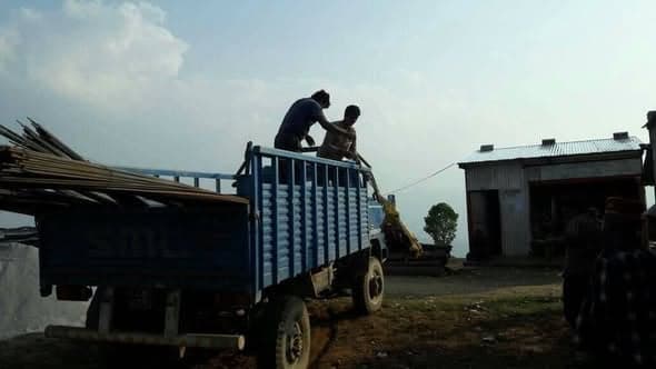 Two men lift bundles of corrugated sheeting into a blue truck outside a workshop, timber stacked nearby.