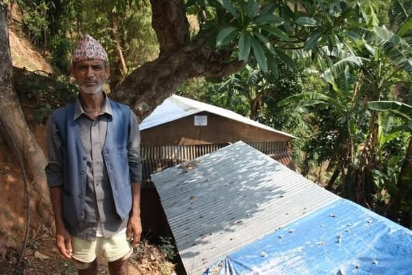 An elderly man in a topi stands on a wooded slope beside two shelters roofed with corrugated iron and tarpaulin.