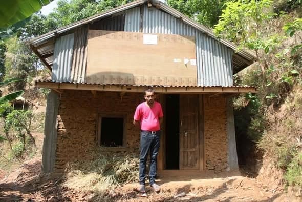 A man in a pink shirt stands in the doorway of a mud house with a plywood gable and corrugated roof.