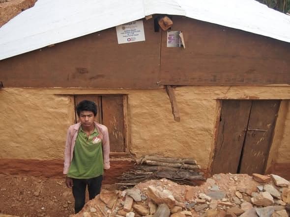 A young man in a green and pink top stands in front of a mud-plastered house with a whitewashed gable and a rough stone yard.