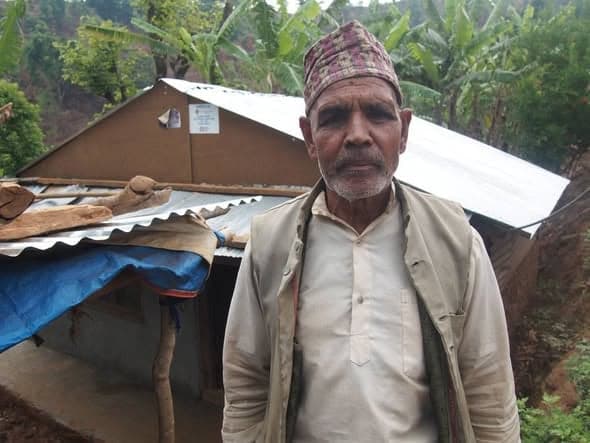 An elderly man in a topi and waistcoat stands in front of a shelter roofed with white sheeting and tarpaulin.