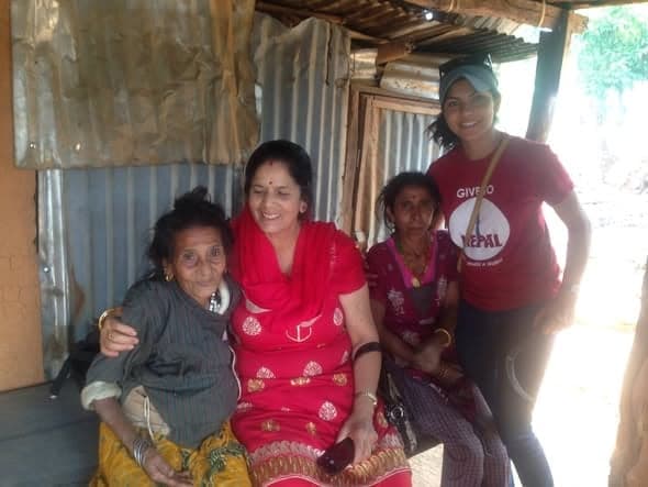 Four women sit and stand together under the corrugated roof of a shelter, one of them in a red Give to Nepal T-shirt.