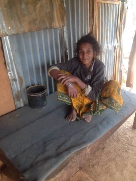An older woman with a bandaged arm sits on a low wooden platform inside a corrugated-iron shelter.