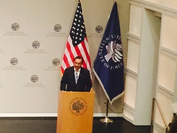 A man in a suit speaks from a lectern between the United States flag and the flag of the United States Institute of Peace.