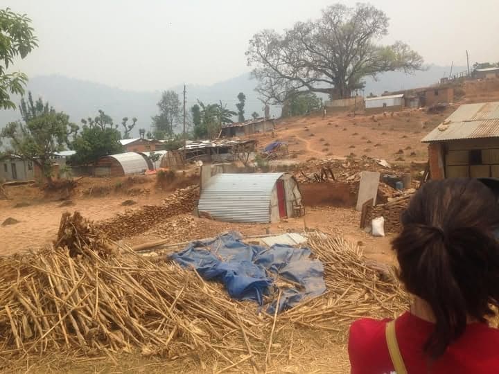 A woman looks out over a hillside settlement of tin shelters, rubble walls and stacked maize stalks under a hazy sky.