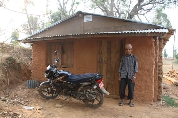 An elderly man stands beside a motorcycle in front of a mud house with a corrugated roof, cut into the hillside.