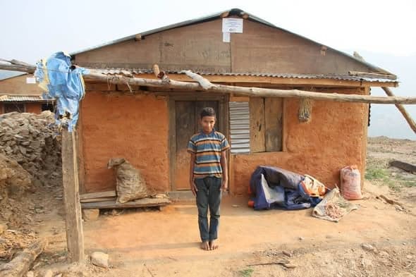 A barefoot boy in a striped shirt stands in the doorway of a mud house, sacks and a tarpaulin bundle stacked against the wall.
