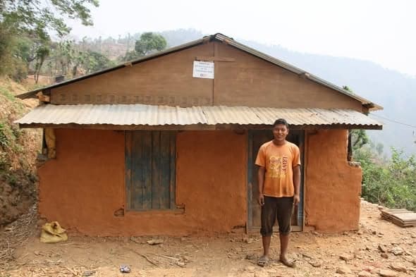 A young man in an orange T-shirt stands in the doorway of a mud house with a blue shuttered window.