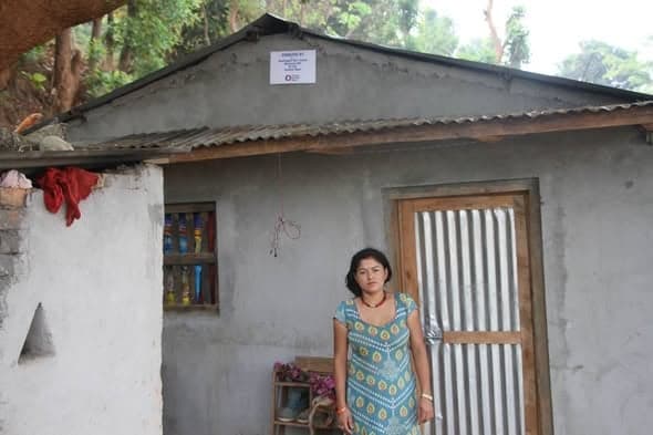 A woman stands beside the doorway of a small cement-rendered house, household goods lined up on a shelf inside the window.