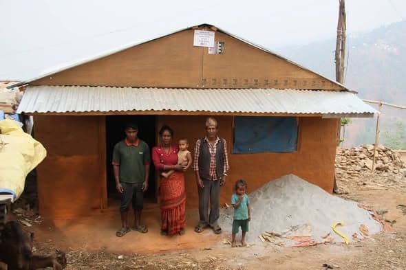 A family of five, including a baby and a small child, stand in front of a mud house with a heap of sand at the door.