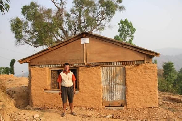 A man in a white and red shirt stands in front of a mud house with a stone-lined window and corrugated roof.