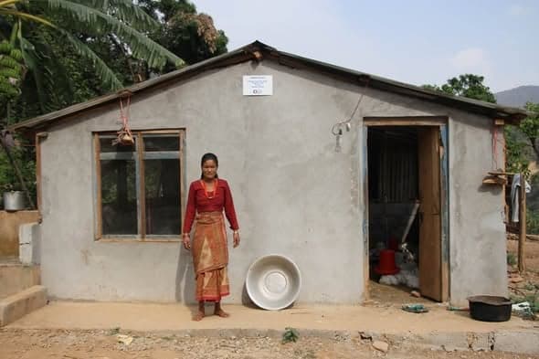 A young woman stands beside a large enamel basin outside a cement-rendered house with a timber window frame.