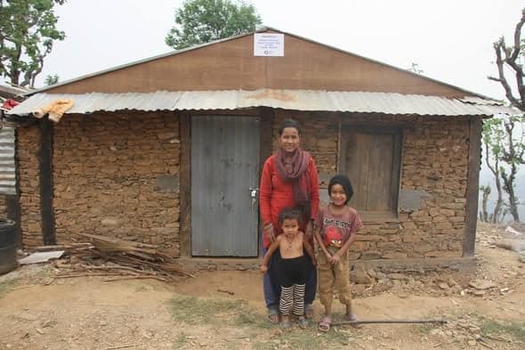 A woman and two young children stand in front of a stone house with a corrugated roof on a bare hilltop.