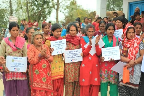 A crowd of women in bright shawls applauding, several of them holding printed cards headed Donated By