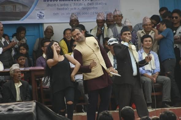 A singer and a dancer perform on a stage in front of a seated audience of villagers and children.