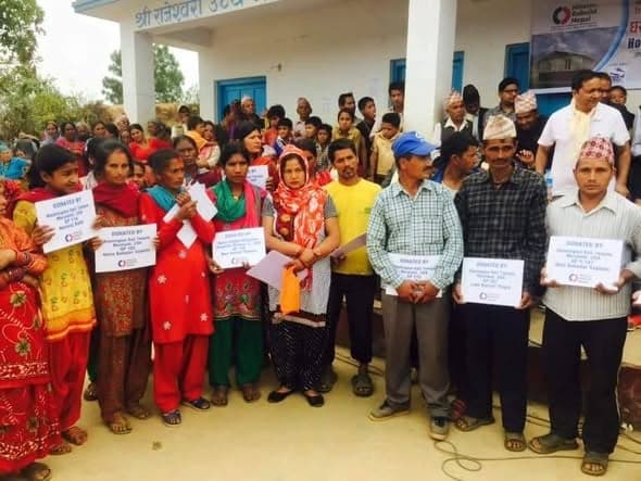 Families standing outside a school building holding printed cards naming the donors who funded their houses.