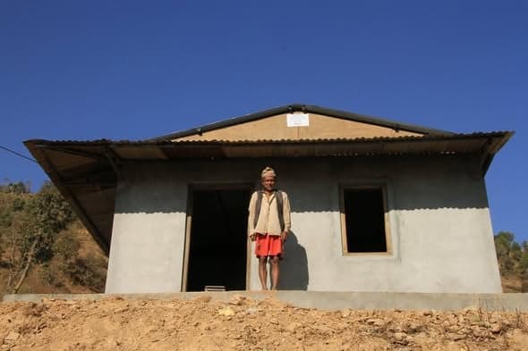An elderly man in a topi standing in the open doorway of a finished single-storey house with plastered walls and a corrugated metal roof, a small plaque fixed above the door.